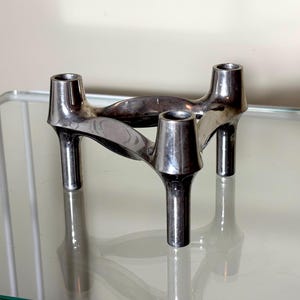 Peut inclure: Un bougeoir en métal argenté avec trois ouvertures cylindriques pour les bougies. Le bougeoir présente un design incurvé unique avec trois pieds. Il est présenté sur une surface en verre.