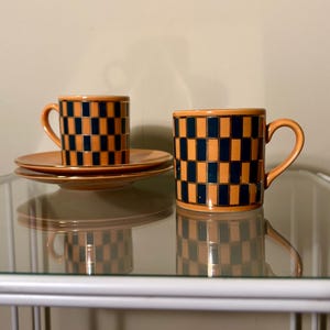 May include: Two orange ceramic coffee mugs and saucers with a checkered pattern of orange and navy blue squares. The mugs have curved handles and are displayed on a glass shelf. The set is a decorative item.