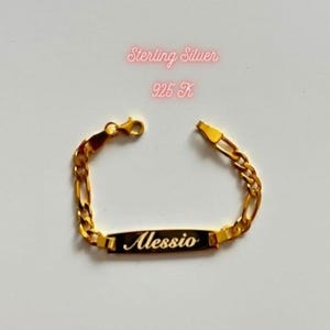 May include: A gold-toned chain bracelet featuring a central rectangular identification plate engraved with the name Alessio in a cursive script. The text Sterling Silver 925 K is visible above the bracelet, which includes a lobster claw clasp.
