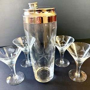 May include: A vintage cocktail shaker with a silver lid and rim, accompanied by five etched martini glasses. The clear glass shaker and glasses feature a wheat stalk design. The set is ideal for serving cocktails.
