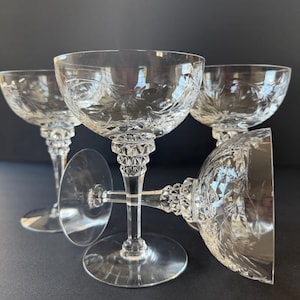 May include: Four clear crystal champagne coupe glasses. The glasses have a round bowl shape with etched floral designs and sit atop a decorative stem. The stems have a diamond pattern. The glasses are arranged on a dark surface.