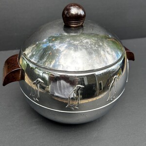May include: A silver-colored ice bucket with a lid and two handles. The lid has a dark brown knob. The bucket has three embossed penguin designs around the base. The handles are dark brown. The bucket reflects the surrounding environment.