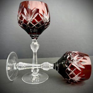 May include: Two wine glasses featuring deep red bowls with intricate diamond and starburst cut patterns. The stems are clear glass with a textured, faceted knop detail, and each glass rests on a wide, circular, clear glass base.