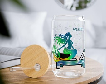 May include: A clear glass water bottle with a bamboo lid and a straw. The bottle has a mermaid design with the word "PILATES" on it.