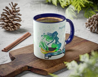May include: White ceramic coffee mug with a blue rim and handle. The mug features a colorful illustration of a mermaid on a pilates reformer with the text "Pilates Mermaid" on the side.