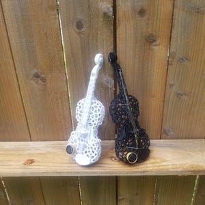 May include: Two decorative, intricately designed violins, one white and one black, are displayed on a wooden shelf. The violins have a detailed, lacy pattern and are positioned side-by-side.