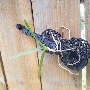 May include: A black, 3D printed, violin-shaped planter with a green plant growing out of it. The planter is attached to a wooden fence.