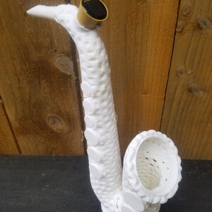 May include: White ceramic saxophone sculpture with a solar panel on top. The sculpture has a textured surface and a hollow interior.
