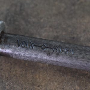 Pu&ograve; includere: Primo piano di un affilacoltelli in acciaio con manico in legno. Il manico &egrave; marrone chiaro e l'asta in acciaio reca la scritta "VOLK STAHL". L'affilacoltelli ha una protezione in metallo argentato.