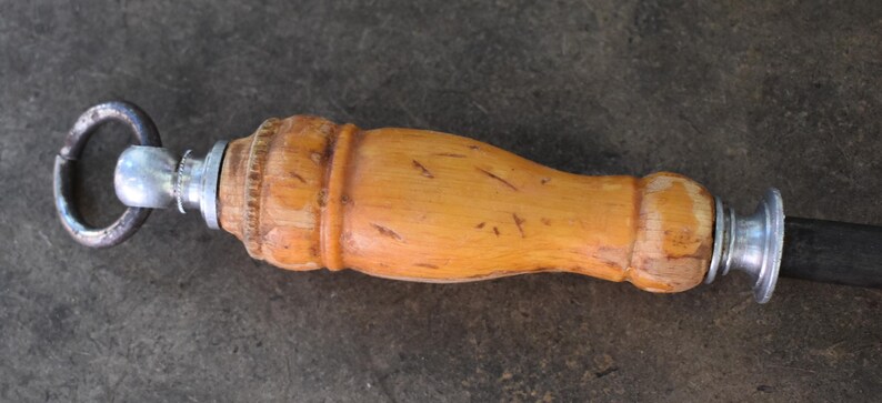 Pu&ograve; includere: Primo piano di un affilacoltelli. L'impugnatura &egrave; in legno chiaro con una forma curva. I componenti metallici sono argentati e l'asta di affilatura &egrave; nera. Un anello di metallo &egrave; attaccato all'estremit&agrave; dell'impugnatura.