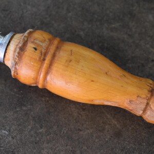 Pu&ograve; includere: Primo piano di un affilacoltelli con manico in legno marrone chiaro e anello metallico. L'impugnatura &egrave; curva e attaccata a un'asta di metallo. Strumento da cucina per affilare i coltelli.