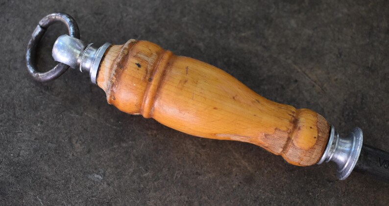 Pu&ograve; includere: Primo piano di un affilacoltelli con manico in legno marrone chiaro e anello metallico. L'impugnatura &egrave; curva e attaccata a un'asta di metallo. Strumento da cucina per affilare i coltelli.