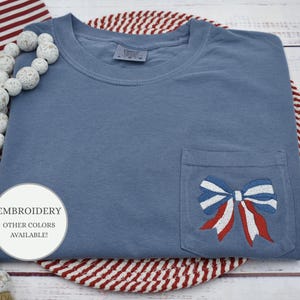 May include: A blue t-shirt with a red, white, and blue bow embroidered on the pocket. The text "EMBROIDERY OTHER COLORS AVAILABLE!" is on a white circle sticker.