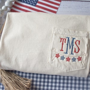 May include: Cream-colored fabric with a pocket embroidered with the initials "TMS" in red and blue, and a garland of red and blue stars. The design has an American patriotic theme.
