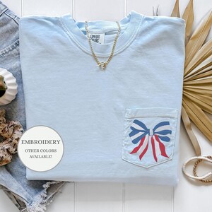 Pode incluir: Camiseta azul claro com um la&ccedil;o vermelho, branco e azul bordado no bolso. O texto "EMBROIDERY OTHER COLORS AVAILABLE!" est&aacute; em um c&iacute;rculo branco.