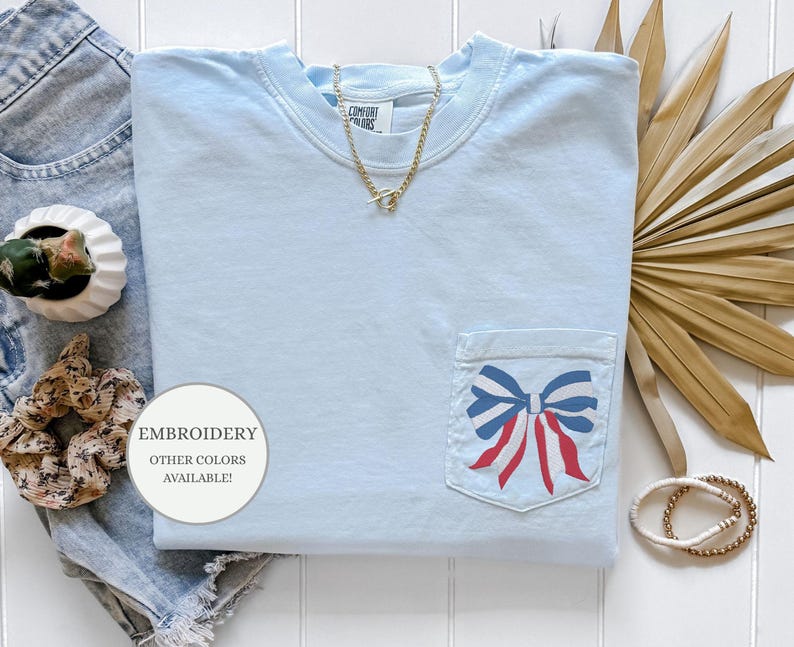 Pode incluir: Camiseta azul claro com um la&ccedil;o vermelho, branco e azul bordado no bolso. O texto "EMBROIDERY OTHER COLORS AVAILABLE!" est&aacute; em um c&iacute;rculo branco.