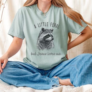 May include: A light blue t-shirt with a black and white illustration of a raccoon and the text "A little feral but Jesus loves me".