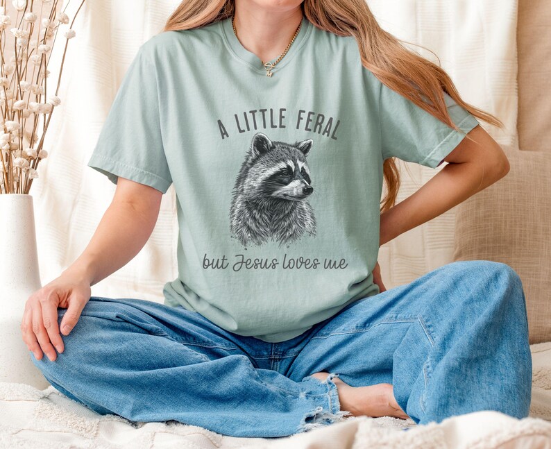 May include: A light blue t-shirt with a black and white illustration of a raccoon and the text "A little feral but Jesus loves me".