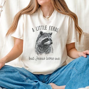 May include: A white t-shirt with a black and white illustration of a raccoon and the text "A little feral but Jesus loves me".