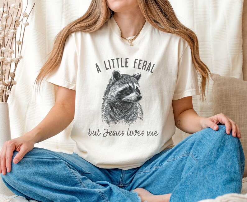 May include: A white t-shirt with a black and white illustration of a raccoon and the text "A little feral but Jesus loves me".
