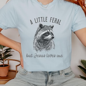 May include: A light blue t-shirt with a black and white illustration of a raccoon. The text "A little feral but Jesus loves me" is printed on the shirt.