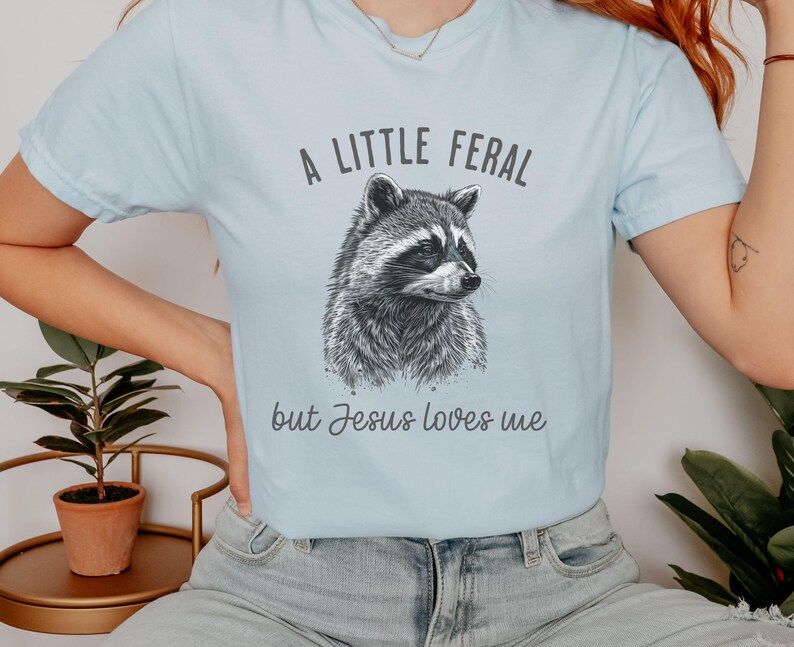 May include: A light blue t-shirt with a black and white illustration of a raccoon. The text "A little feral but Jesus loves me" is printed on the shirt.