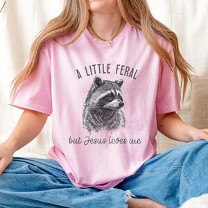 May include: A pink t-shirt with a black and white illustration of a raccoon and the text "A little feral but Jesus loves me".