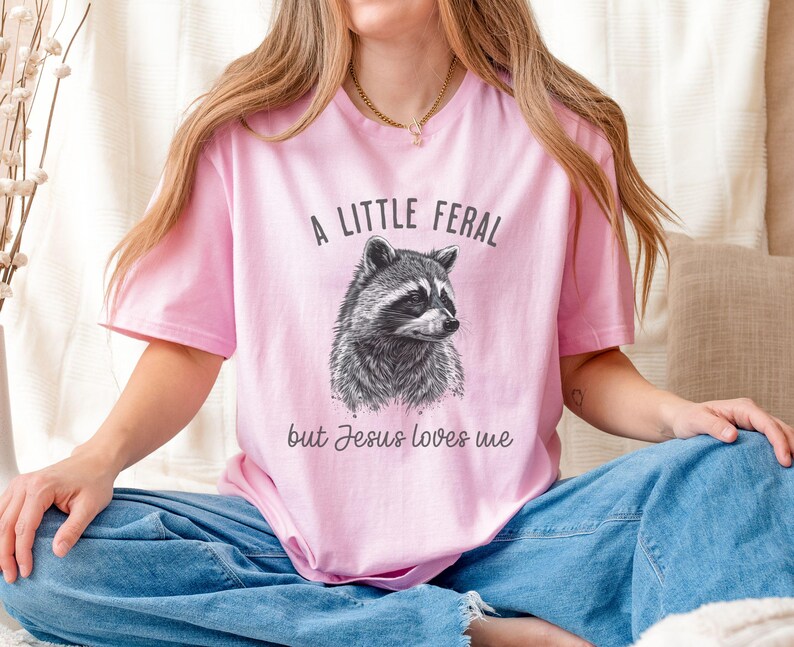 May include: A pink t-shirt with a black and white illustration of a raccoon and the text "A little feral but Jesus loves me".