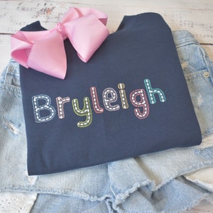 May include: A navy blue sweatshirt with the name "Bryleigh" embroidered in colorful thread on the front. The sweatshirt is folded and placed on top of a pair of light blue denim shorts.