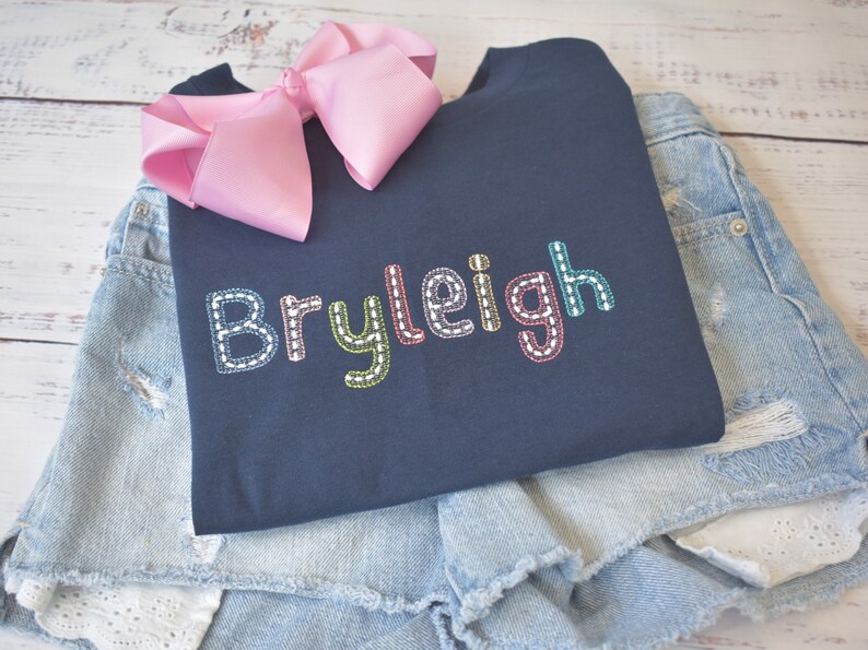 May include: A navy blue sweatshirt with the name "Bryleigh" embroidered in colorful thread on the front. The sweatshirt is folded and placed on top of a pair of light blue denim shorts.