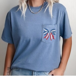 Pode incluir: Uma camiseta azul claro com um la&ccedil;o vermelho, branco e azul em um bolso.