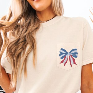 Pode incluir: Uma camiseta bege com um la&ccedil;o vermelho, branco e azul no bolso.