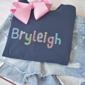 May include: A navy blue t-shirt with the name "Bryleigh" embroidered in colorful thread. The shirt is folded and placed on top of a pair of light blue denim shorts.