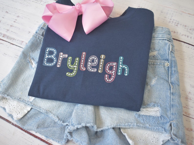 May include: A navy blue t-shirt with the name "Bryleigh" embroidered in colorful thread. The shirt is folded and placed on top of a pair of light blue denim shorts.