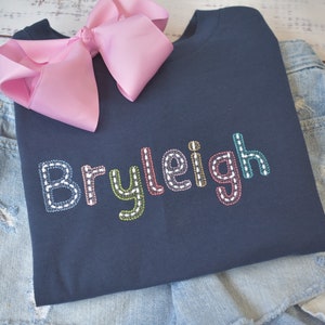 May include: A navy blue t-shirt with the name "Bryleigh" embroidered in colorful thread. The letters are outlined in white and have a stitched look.