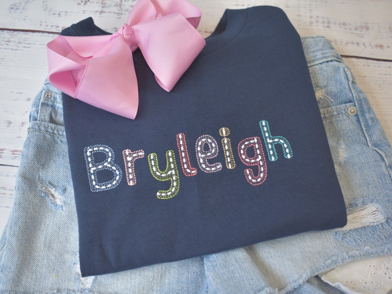 May include: A navy blue t-shirt with the name "Bryleigh" embroidered in colorful thread. The letters are outlined in white and have a stitched look.