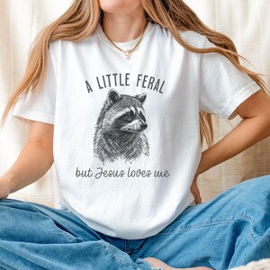 May include: White t-shirt with a black and white illustration of a raccoon and the text "A little feral but Jesus loves me."