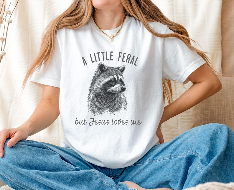 May include: White t-shirt with a black and white illustration of a raccoon and the text "A little feral but Jesus loves me."