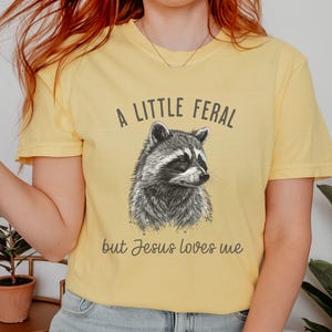 May include: A yellow t-shirt with a black and white illustration of a raccoon and the text "A little feral but Jesus loves me".