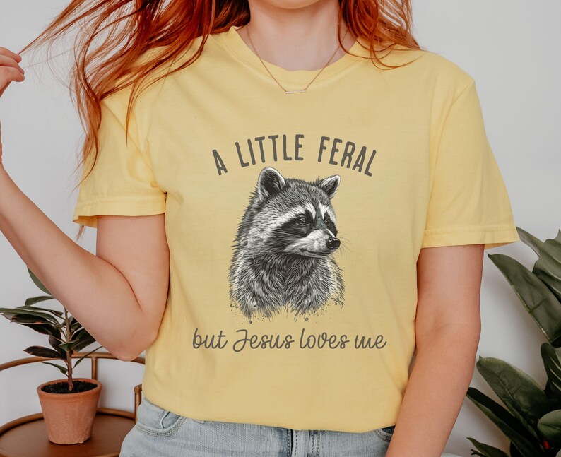 May include: A yellow t-shirt with a black and white illustration of a raccoon and the text "A little feral but Jesus loves me".