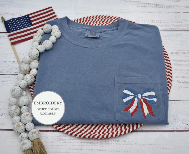May include: A blue t-shirt with a red, white, and blue bow embroidered on the pocket. The text "EMBROIDERY OTHER COLORS AVAILABLE!" is on a white circle sticker.