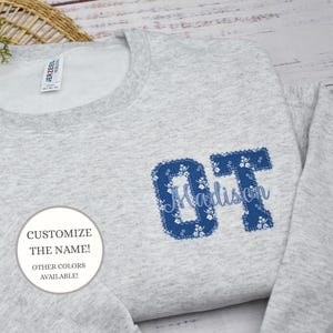 May include: A light gray sweatshirt with the letters "OT" in a blue floral pattern, with the name "Madison" in a light blue script. The image also includes a white circle with the text "CUSTOMIZE THE NAME! OTHER COLORS AVAILABLE!"