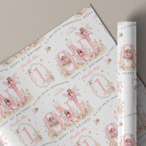May include: White wrapping paper featuring a repeating pattern of pink fairy illustrations, floral accents, and the text "HAPPY 1ST BIRTHDAY TO A VERY SPECIAL GRANDDAUGHTER" and "Amelia Mai" alongside large pink numbers and fairy motifs.