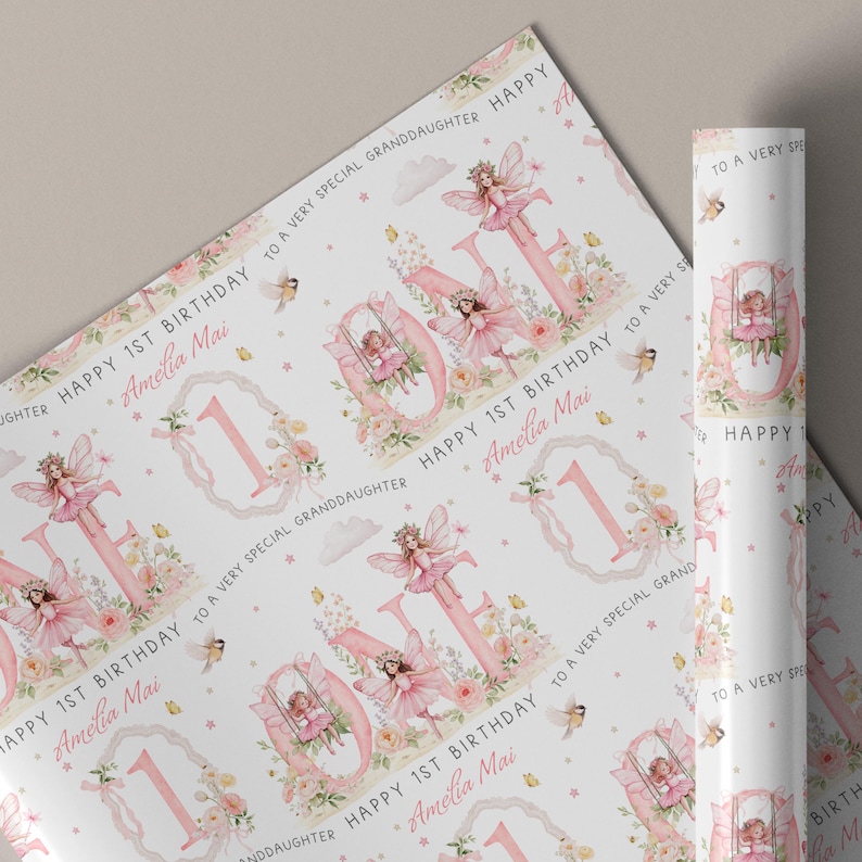 May include: White wrapping paper featuring a repeating pattern of pink fairy illustrations, floral accents, and the text "HAPPY 1ST BIRTHDAY TO A VERY SPECIAL GRANDDAUGHTER" and "Amelia Mai" alongside large pink numbers and fairy motifs.