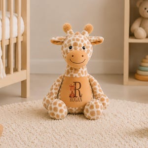 May include: A plush giraffe toy with tan and white spotted fur, featuring a solid tan belly patch printed with a small giraffe illustration, the letter R, and the name ROGAN. The stuffed animal has two small horns and a stitched smiling face.