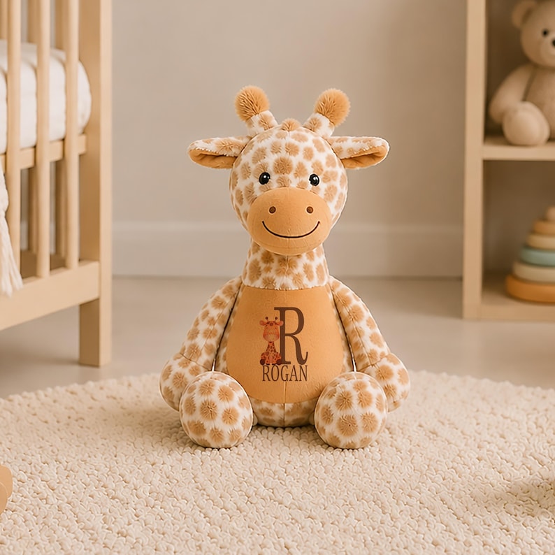 May include: A plush giraffe toy with tan and white spotted fur, featuring a solid tan belly patch printed with a small giraffe illustration, the letter R, and the name ROGAN. The stuffed animal has two small horns and a stitched smiling face.