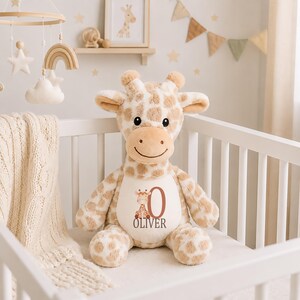 May include: A plush giraffe toy with tan and white spots, featuring a cream-colored belly patch printed with a small giraffe illustration, the large letter O, and the name OLIVER in dark brown text. The toy has soft fur, small horns, and ears.