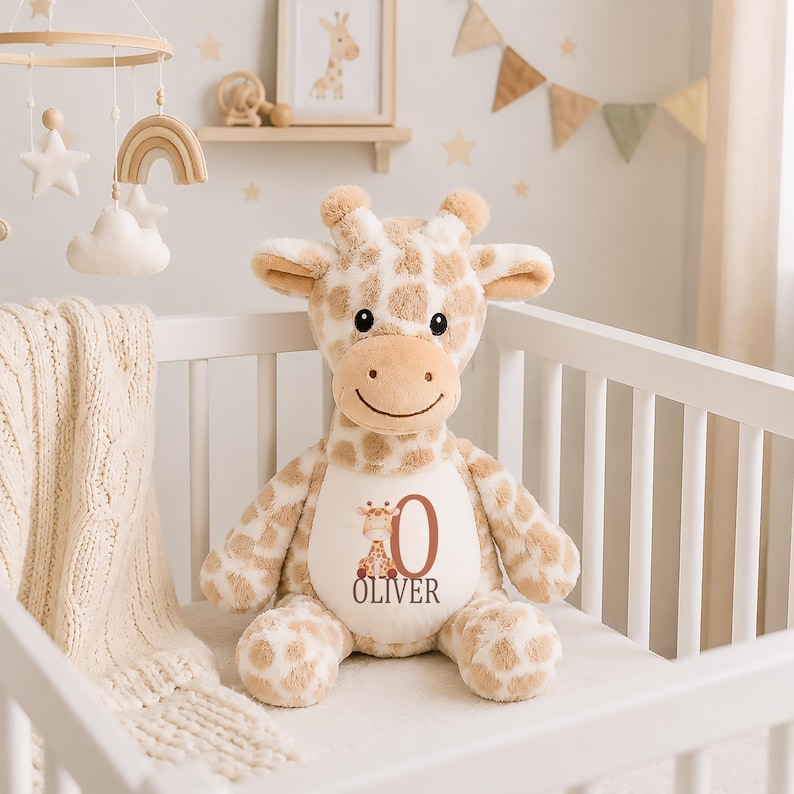 May include: A plush giraffe toy with tan and white spots, featuring a cream-colored belly patch printed with a small giraffe illustration, the large letter O, and the name OLIVER in dark brown text. The toy has soft fur, small horns, and ears.