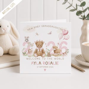 May include: A white card with the text "NEW BABY GRANDDAUGHTER" and "WELCOME TO THE WORLD". The card features illustrations of a teddy bear, lamb, and other baby-themed graphics. The name "AYLA ROSALIE" and the date "21 SEPTEMBER 2026" are also displayed.