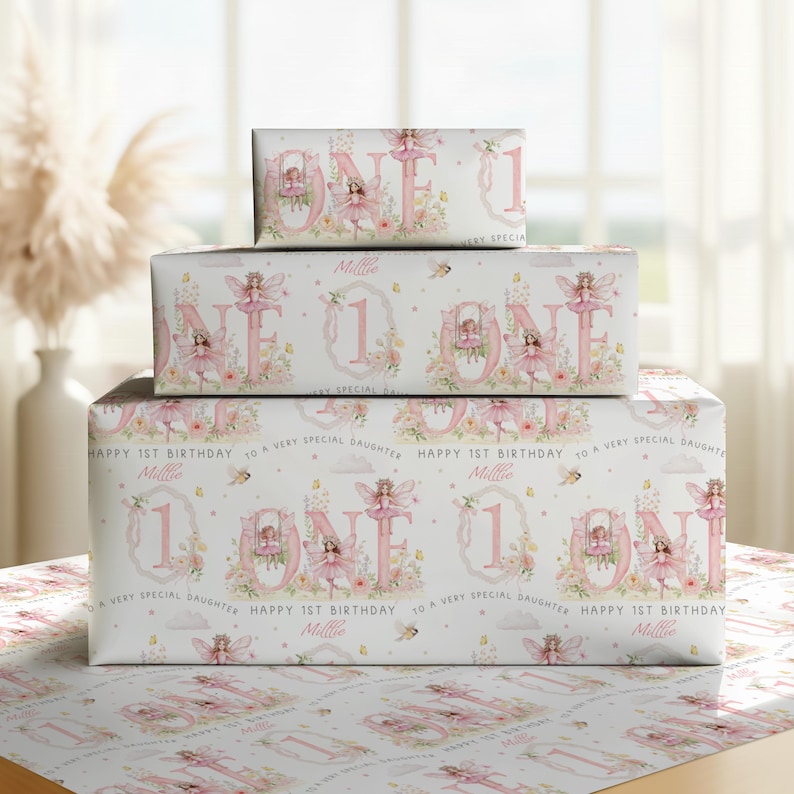 May include: Three stacked rectangular gift boxes wrapped in white paper featuring a repeating pattern of pink fairies, floral accents, and the text "ONE" and "HAPPY 1ST BIRTHDAY TO A VERY SPECIAL DAUGHTER" with the name "Millie" printed in pink.
